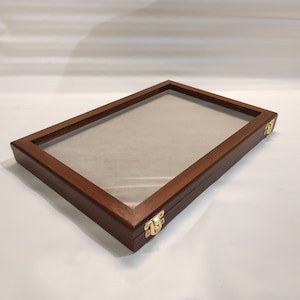 Puede incluir: Una vitrina de madera con tapa de cristal y cierres de latón. La vitrina está vacía y tiene un acabado marrón claro.