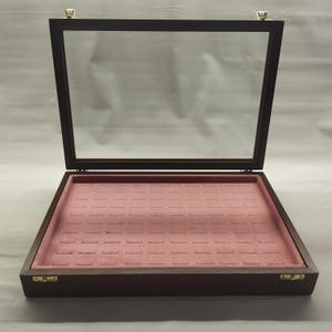Könnte beinhalten: Offene Holzvitrine mit Glasdeckel. Der Innenraum verfügt über einen rosa Samteinsatz mit mehreren Schlitzen. Das Gehäuse hat eine dunkelbraune Oberfläche und messingfarbene Scharniere und Verschlüsse. Ideal für die Präsentation von Schmuck oder kleinen Sammlerstücken.