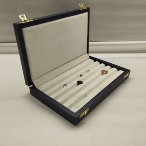 May include: A black velvet jewellery box with a hinged lid and gold clasps. The box is open and has multiple compartments for storing small items, such as gemstones. The compartments are lined with white velvet.