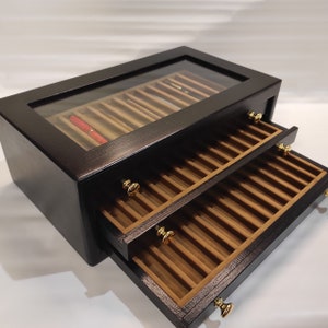 Könnte beinhalten: Schwarze Holzstiftebox mit Glasdeckel und zwei Schubladen. Die Schubladen sind mit braunem Filz ausgekleidet und haben goldfarbene Knöpfe. Die Box ist für Stifte und andere Schreibutensilien konzipiert.