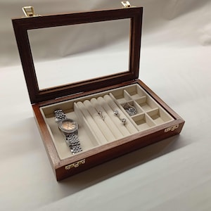 May include: Open wooden jewelry box with a clear glass lid. Inside, a silver wristwatch with a rose-gold face and several rings are displayed on a cream-colored velvet lining. The box is made of dark wood.