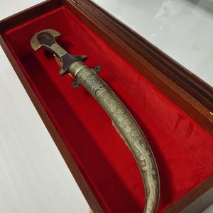 May include: An ornate dagger with a curved blade, housed in a wooden display case lined with red velvet. The dagger features a silver-colored handle and blade with intricate engraved patterns. The handle is dark brown.