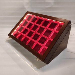 Könnte beinhalten: Eine Holzauslage mit Glasdeckel und roter Samtfütterung. Die Auslage hat 24 Fächer und wird durch LED-Leuchten beleuchtet.