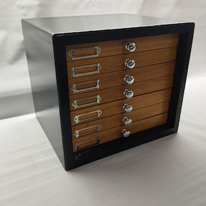May include: A wooden cabinet with six drawers. The cabinet has a black frame and a light-coloured top. Each drawer has a silver label holder and a round, silver knob. The drawers are made of wood with a visible grain.