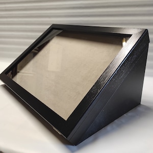 May include: A black wooden display case with a glass front and a beige felt lining. The case is angled and has a beveled edge.