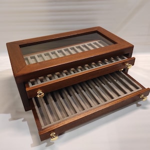 Könnte beinhalten: Eine Holzstiftebox mit Glasdeckel und zwei Schubladen. Die Schubladen sind geöffnet und haben Fächer für Stifte. Die Box ist aus dunklem Holz mit Messingakzenten.