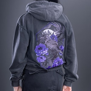 Puede incluir: Una sudadera con capucha gris carbón que presenta un gran gráfico en la espalda. El diseño muestra un cuervo negro detallado posado entre flores y follaje de color púrpura vibrante, sobre una luna circular y un marco geométrico oscuro.
