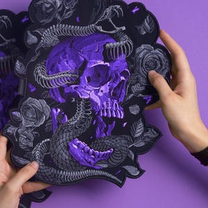 Puede incluir: Dos manteles individuales negros con un diseño de calavera y serpiente morada. La calavera está en el centro, con una serpiente enrollada alrededor. También se presentan rosas y hojas grises. Los manteles individuales se sostienen sobre un fondo morado.