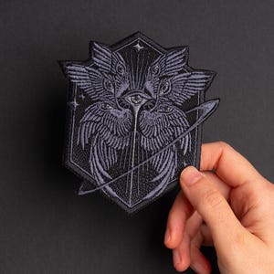 Könnte beinhalten: Ein schwarz-grauer, gestickter Aufnäher mit einem stilisierten Engel-Design mit mehreren Augen und Flügeln. Der Aufnäher hat die Form eines Schildes mit einem Stern oben und einer gebogenen Linie um die zentrale Figur.