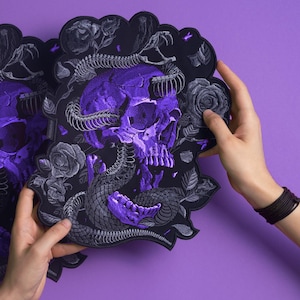 May include: A large, die-cut sticker or patch featuring a vibrant purple human skull entwined with a detailed grey snake skeleton. The design is surrounded by intricate grey rose illustrations on a black background, creating a high-contrast visual.