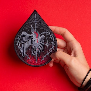 May include: A black, teardrop-shaped patch featuring a stylized bird pierced by a sword. The bird is gray with red accents, surrounded by thorny vines and stars. The patch is held against a red background.