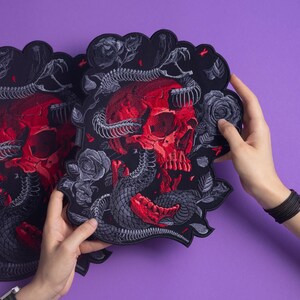 以下が含まれることがあります： 黒い刺繍ワッペン。赤い頭蓋骨に灰色の骸骨の蛇が絡みつき、周囲を灰色のバラが囲むデザイン。黒い背景にスカラップ状の縁取りが施され、精巧な刺繍糸の質感が特徴的です。