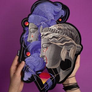 Może przedstawiać: Haftowana naszywka przedstawiająca dwa klasyczne popiersia kobiet z płynącymi włosami i łzami. Projekt zawiera geometryczne kształty, czerwone i żółte akcenty oraz tekst "DAMONK". Naszywka znajduje się na czarnym tle z fioletowym tłem.