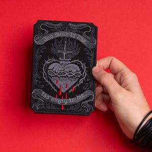 May include: A black embroidered patch featuring a sacred heart design with a crown of thorns, a sword, and dripping blood. The patch has the phrases "Too mean to live" and "Too angry to die" in a banner design.