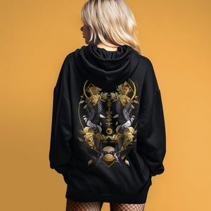 Könnte beinhalten: Schwarzer Kapuzenpullover mit einem detaillierten gold-schwarzen Grafikdesign auf dem Rücken. Das Design zeigt zwei Vögel, Rosen und geometrische Formen. Der Hoodie hat eine lockere Passform und eine Kapuze.