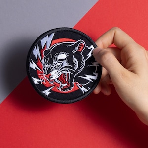 Può includere: Toppa ricamata in bianco e nero di una pantera su sfondo rosso con fulmini bianchi. La toppa è su uno sfondo nero.