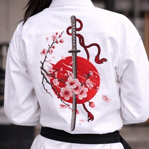Puede incluir: Uniforme blanco de artes marciales con cintur&oacute;n negro. La espalda del uniforme presenta un dise&ntilde;o bordado de una espada katana, un sol rojo y flores de cerezo. El dise&ntilde;o es detallado y colorido.