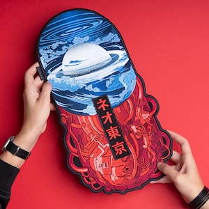 May include: Embroidered patch featuring a celestial design with a planet and swirling blue clouds above a red, mechanical-looking lower section. The patch includes Japanese text. The patch is held against a red background.