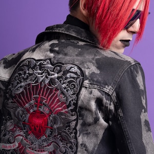 Op de afbeelding: Zwart denim jack met een grote geborduurde patch op de rug. De patch heeft een hart met vleugels, een kroon en een banner met de tekst "Ruled by Passion".