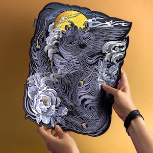 Può includere: Toppa ricamata con un design dettagliato di una creatura mitica, luna dorata ed elementi floreali. La toppa è principalmente nera, grigia e bianca, con accenti dorati. La toppa è tenuta da due mani. Altezza circa 30 cm.