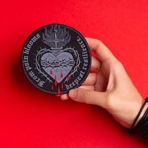 May include: A round, black patch featuring a gray heart pierced by a sword, surrounded by a crown of thorns. Red blood drips from the heart. The text "From pain blooms deepest resilience" is arched around the design.