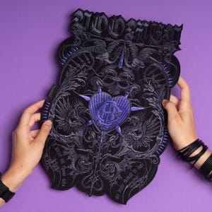 May include: A black and purple embroidered patch with the word "RIDORFUL" at the top. The patch features a heart design with an "R" in the centre, surrounded by ornate details and text. The patch is held against a purple background.