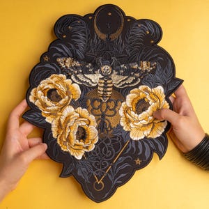 May include: A large, black, embroidered patch featuring a central moth with detailed wings, surrounded by three prominent yellow and white peony flowers. The design includes intricate dark grey foliage, a crescent moon, and a thin golden line.