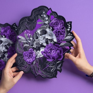 May include: An embroidered patch featuring a detailed grayscale human skull surrounded by vibrant purple peony flowers and delicate white floral sprigs. The design includes intricate black thread work and a purple geometric background element.