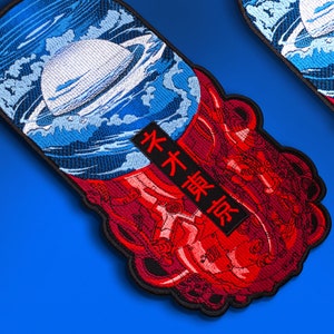 May include: Embroidered patch featuring a planet with rings surrounded by blue waves and red, stylized tentacles. A black rectangle with Japanese text is also present. The patch has a black border and is set against a blue background.
