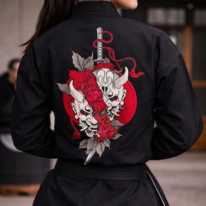 Op de afbeelding: Zwart vechtsportuniform met een gedetailleerd geborduurd ontwerp op de rug. Het ontwerp toont twee witte demonenmaskers, rode bloemen, een zwaard en een rode cirkel. Het uniform heeft een zwarte riem.