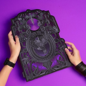 Puede incluir: Un objeto decorativo rectangular con un diseño gris oscuro detallado sobre un fondo púrpura. El diseño presenta una imagen central de un pájaro dentro de un círculo, rodeada de patrones y figuras ornamentadas. El objeto está sostenido por dos manos.