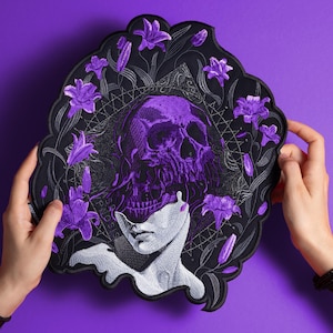 May include: A black patch with a purple skull design, surrounded by purple flowers and a woman's face. The patch has the words "MASS HYPNOSIS" in white. The patch is held against a purple background.