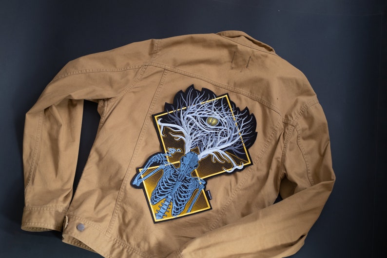 May include: A tan jacket with a large, detailed patch on the back. The patch features a stylized design of a skeleton with a tree-like structure and an eye, in shades of blue, white, and yellow. The jacket is made of a durable fabric.