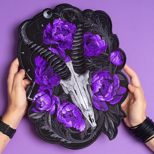 May include: A black embroidered patch featuring a goat skull with large, curved horns, surrounded by vibrant purple peony flowers. The design includes a crescent moon and decorative flourishes, creating a gothic floral aesthetic.