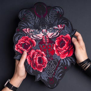 May include: A large, shaped embroidered patch featuring a central death's-head hawkmoth with pink and grey wings, surrounded by three vibrant red blooming peonies and intricate dark grey foliage on a black background with fine line details.
