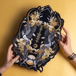 Pode incluir: Patch bordado preto com um desenho de pássaros em voo, rodeados por rosas douradas e folhagens. O patch tem uma borda decorativa e inclui um gráfico de onda sonora. O patch é segurado contra um fundo dourado.