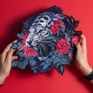 Puede incluir: Parche bordado con la cara de un tigre con rayas blancas y negras, rodeado de rosas rojas y hojas azul-verdosas. El parche tiene un fondo azul oscuro y un borde rojo, creando un contraste llamativo sobre el fondo rojo.