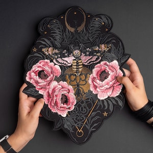 May include: A large, shield-shaped embroidered patch featuring a central moth with pink and black wings, positioned above three detailed pink peony flowers. The design includes gold filigree, a crescent moon, and star accents on a black background.