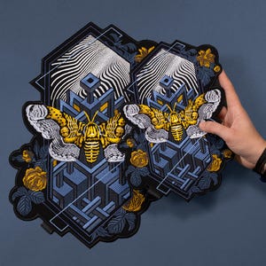 May include: Two embroidered patches with a yellow and black moth design. The patches have a geometric blue and black background, with yellow roses and white wave patterns. The patches are held by a hand. The patches are suitable for clothing.