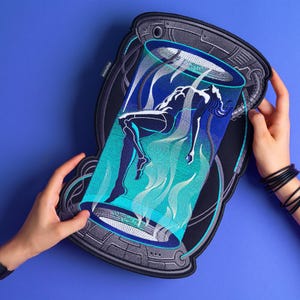 May include: A large, embroidered patch featuring a figure suspended in a glowing blue and teal chamber. The patch has a dark gray border with mechanical details. The figure is a white silhouette against the blue and teal background. The patch is held against a blue background.