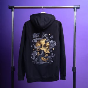 Peut inclure: Un sweat à capuche noir présentant une broderie détaillée au dos représentant un crâne humain doré entrelacé d'un serpent sombre et entouré de roses grises. Le motif inclut de petits accents dorés et des détails floraux complexes sur le tissu.