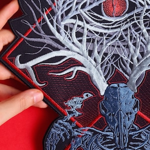 May include: Embroidered patch with a skull, antlers, and eye design. The patch has a black background with red and white detailing. The design is intricate and artistic, suitable for jackets or bags. The patch is approximately 15 cm in length.