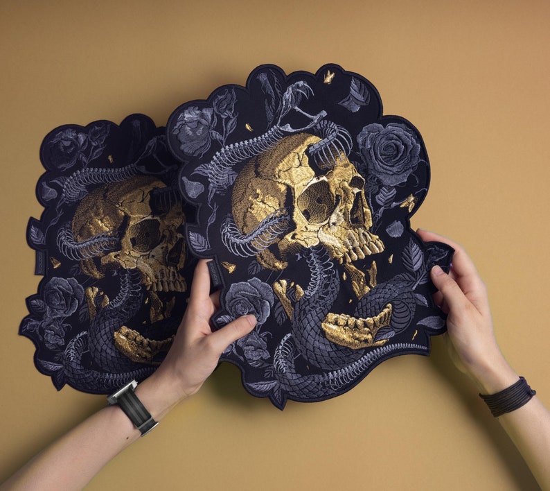 May include: Two black decorative pieces with a gold skull and snake design. The skull is in the center, with a snake coiled around it. Dark roses and leaves surround the skull and snake. The pieces are held up against a tan background.