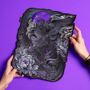 Puede incluir: Un parche bordado rectangular de gran tamaño con un motivo central de fénix en tonos negros y grises, acentuado por una luna púrpura vibrante, nubes arremolinadas y elementos florales detallados con patrones de plumas de pavo real.