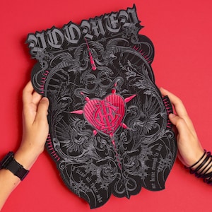 May include: A large, black embroidered patch with a gothic design. The patch features a pink heart in the centre, surrounded by intricate grey details, including dragons and text. The patch is held against a red background.