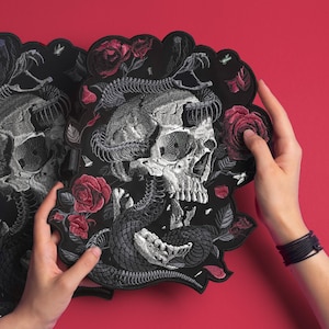 Peut inclure: Écussons noirs brodés avec un motif de crâne, un serpent et des roses rouges. Le crâne est détaillé avec des ombres et des textures, et le serpent l'entoure. Les roses sont en pleine floraison.