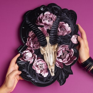 Könnte beinhalten: Schwarze, gestickte Wandkunst mit einem Schädel mit großen, gebogenen Hörnern, umgeben von rosa und lila Pfingstrosen. Das Kunstwerk wird von Händen vor einem magentafarbenen Hintergrund gehalten. Das Design umfasst komplizierte Blumen- und Scrollwork-Details.