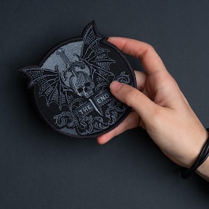 May include: A black and gray embroidered patch featuring a skull with bat wings, a sword, and the words "THE END." The patch has a circular shape with pointed edges at the top. The design is detailed and gothic in style.