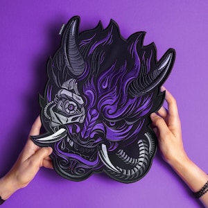 May include: A large embroidered patch featuring a stylized oni mask design in shades of purple, black, and silver. The mask incorporates mechanical elements, including a robotic eye and segmented tubing, with sharp horns and prominent fangs.