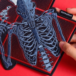 May include: Embroidered patch featuring a blue skeletal design on a black background, framed by a red border. The patch has a diamond shape with a small tag that reads "graphicine". The design includes a detailed depiction of a rib cage and spine.